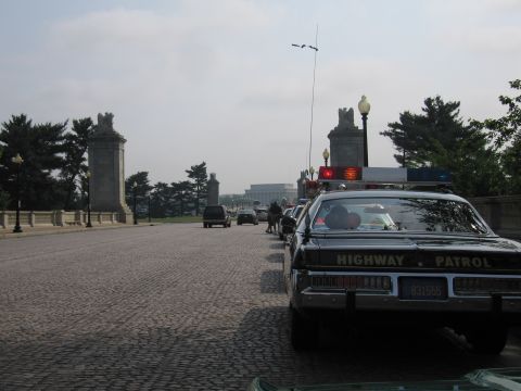 1991 Ford Crown Victoria - A line of vehicles, including a highway patrol car, are stopped on a bridge with large stone structures and trees in the background.