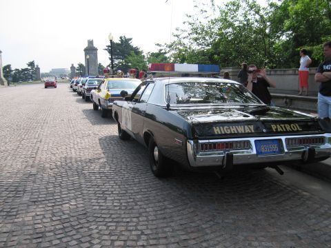 1991 Ford Crown Victoria - A line of vintage police cars, including a black highway patrol vehicle, are parked on a cobblestone street.