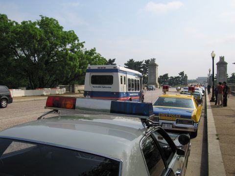 1991 Ford Crown Victoria - Several police cars and a tour bus are lined up on a bridge, with a distant monument visible.