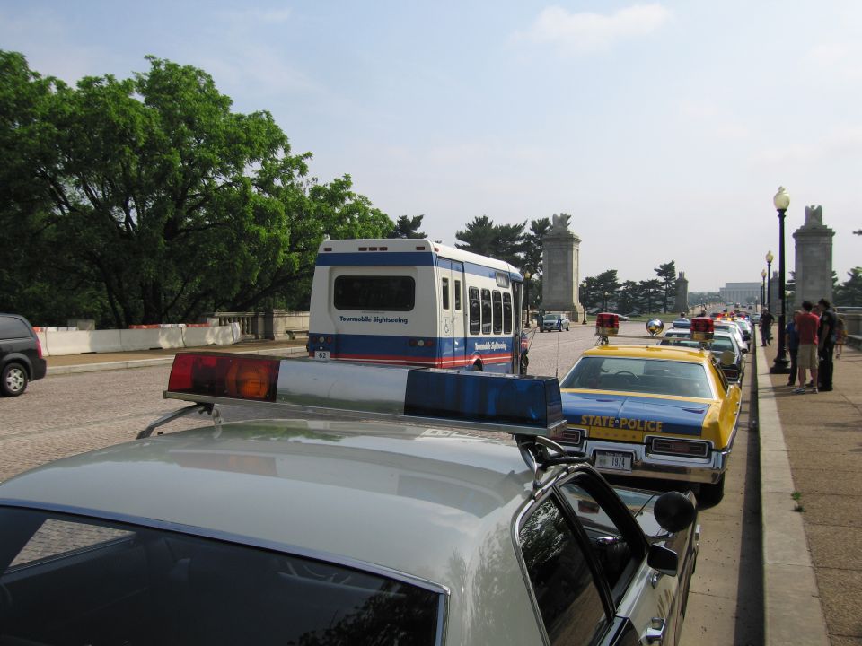 Police cars and a sightseeing bus lined up on a road with a yellow State Police car
