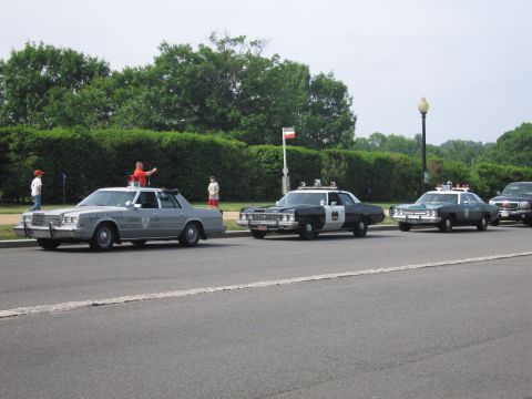 1991 Ford Crown Victoria - Several police vehicles are parked in a row, with a person standing on the hood of the lead car.