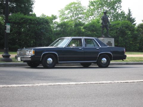 1991 Ford Crown Victoria - A dark sedan is parked on the side of the road, with a statue visible behind it.