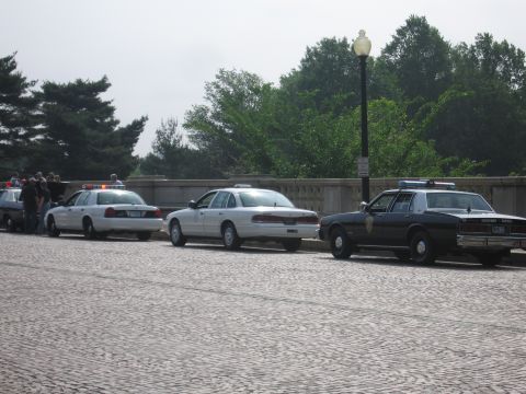 1991 Ford Crown Victoria - Three police cars, two white and one black, are parked in a line on a cobblestone street. The black car has 'HIGHWAY PATROL' text on its si…