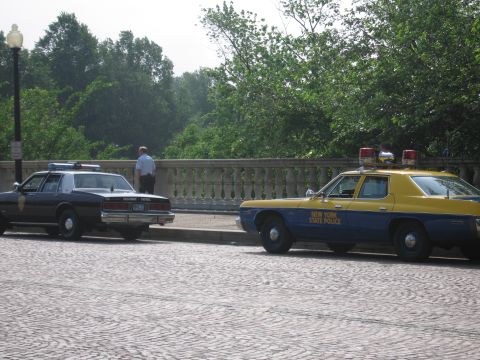 1991 Ford Crown Victoria - Two police cars, one yellow and blue, the other dark, are parked on a cobblestone street.