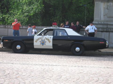 1991 Ford Crown Victoria - A vintage police car with a light bar on top is seen from the side, with several people standing nearby.