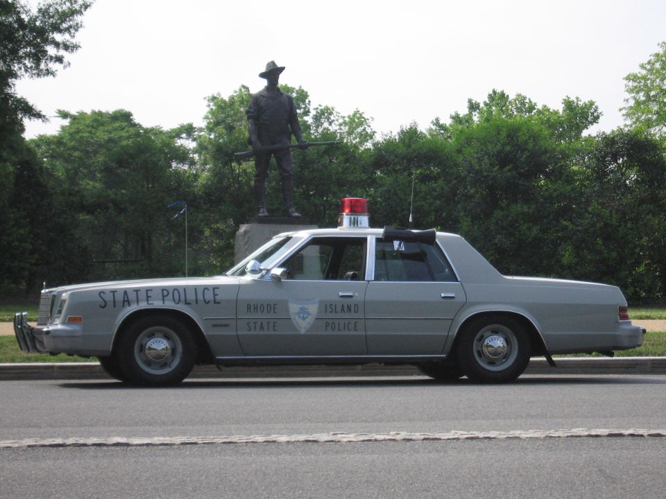 Gray Rhode Island State Police sedan parked on a road in front of a statue
