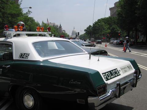 1991 Ford Crown Victoria - A vintage police car with a light bar is parked on a street, with other vehicles and buildings in the background.