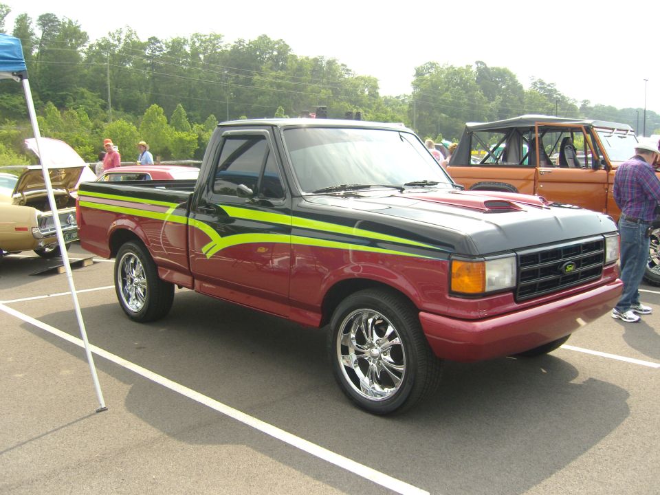 Red and black Ford F-150 pickup with green and yellow graphics, chrome wheels, and a hood scoop. Another truck and car are parked nearby