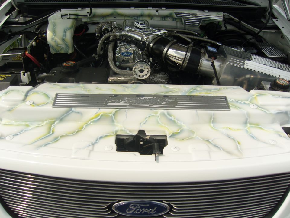 Close-up of a white truck engine bay with custom green and blue marble paint on the engine cover and air intake. Ford logo visible on grille