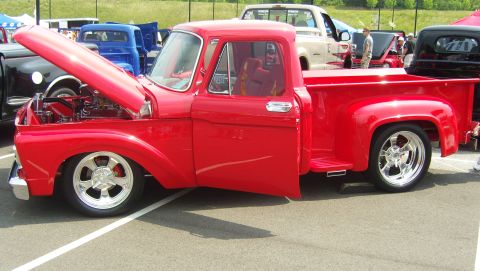 2007 Ford Mustang - Side view of a lowered red 1960s Ford F-100 stepside pickup at an outdoor show with its hood tilted open over a chromed engine and polished…