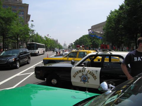 1991 Ford Crown Victoria - Multiple highway patrol vehicles are lined up on a street, with buildings and trees in the background.