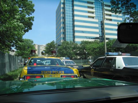 1991 Ford Crown Victoria - Several police vehicles are stopped in traffic on a sunny day, with a modern office building in the background.
