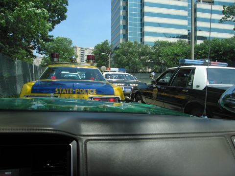 1991 Ford Crown Victoria - A line of police cars, including a yellow and blue state police vehicle, are stopped on a street.