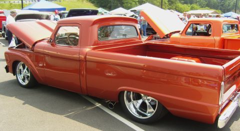 2007 Ford Mustang - A customized orange pickup truck is displayed at an outdoor event with its hood open, parked beside another similar vehicle.