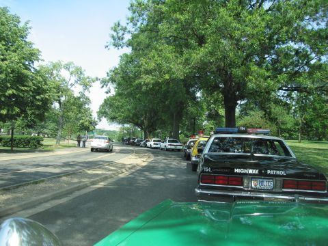1991 Ford Crown Victoria - Several police cars are parked in a line on a road next to a park with trees.