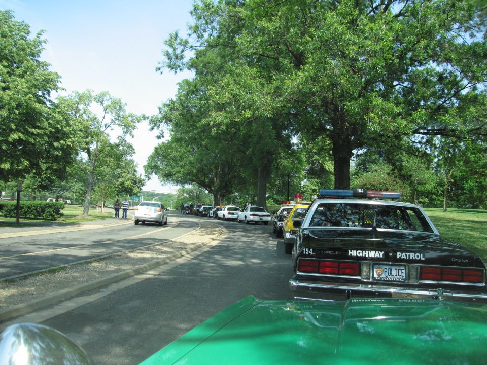 Black Chevrolet Highway Patrol car parked in a line of vehicles under trees