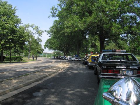 1991 Ford Crown Victoria - Several police vehicles are parked in a line on the side of a road under a canopy of trees.