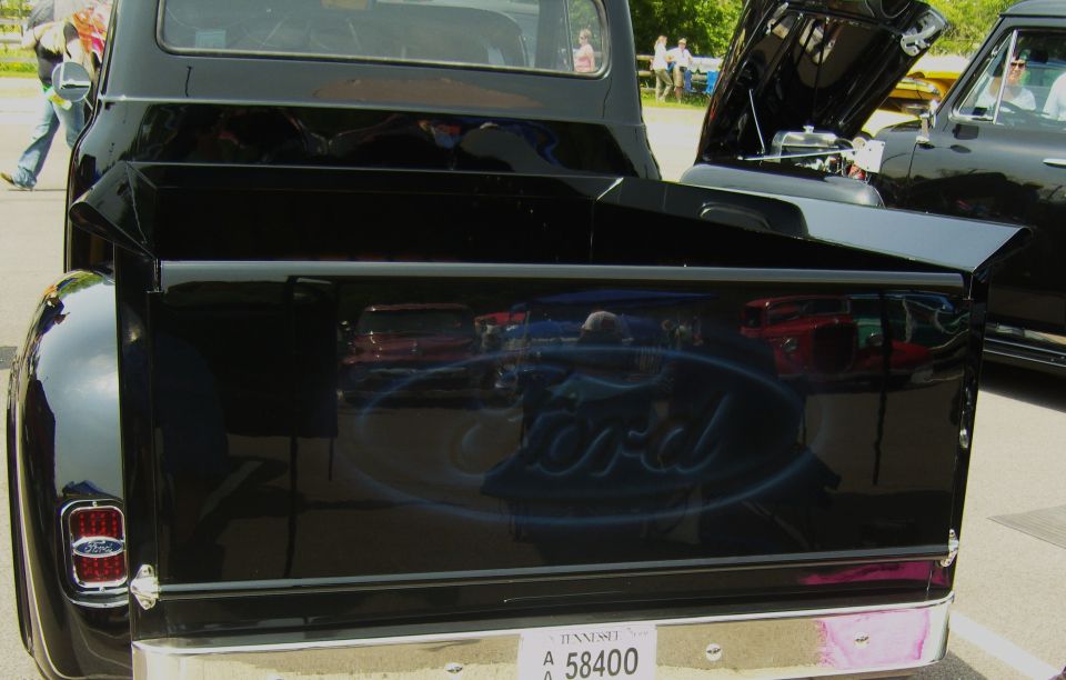 Black Ford pickup truck's tailgate with a blue Ford logo airbrushed on it. Another black vehicle with its hood up is next to it