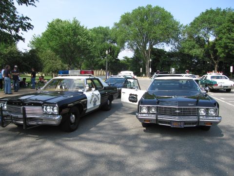 1991 Ford Crown Victoria - Several police cars are parked on a sunny day, with trees and people visible in the background.