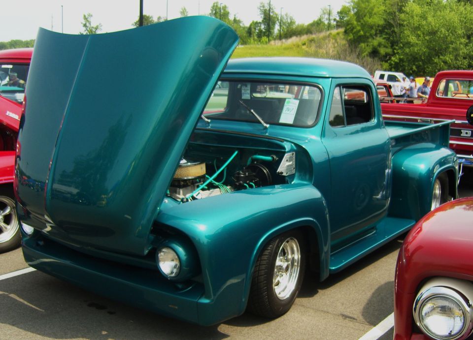 Teal 1956 Ford F-100 pickup truck with hood open, showing engine. Another red truck is partially visible