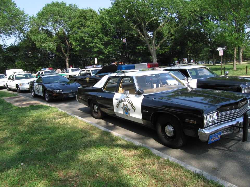 Black-and-white Dodge Highway Patrol sedan parked on grass by a Camaro