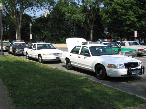 1991 Ford Crown Victoria - A line of police cars, including a white 1991 Ford Crown Victoria with its hood up, are parked on the side of the road.