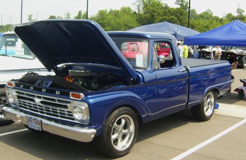 2007 Ford Mustang - A blue 1965 Ford F-100 pickup truck is displayed with its hood open, revealing the engine, at an outdoor event.