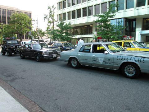 1991 Ford Crown Victoria - A line of police vehicles, including a silver state police car and a black unmarked car, are parked on a city street.