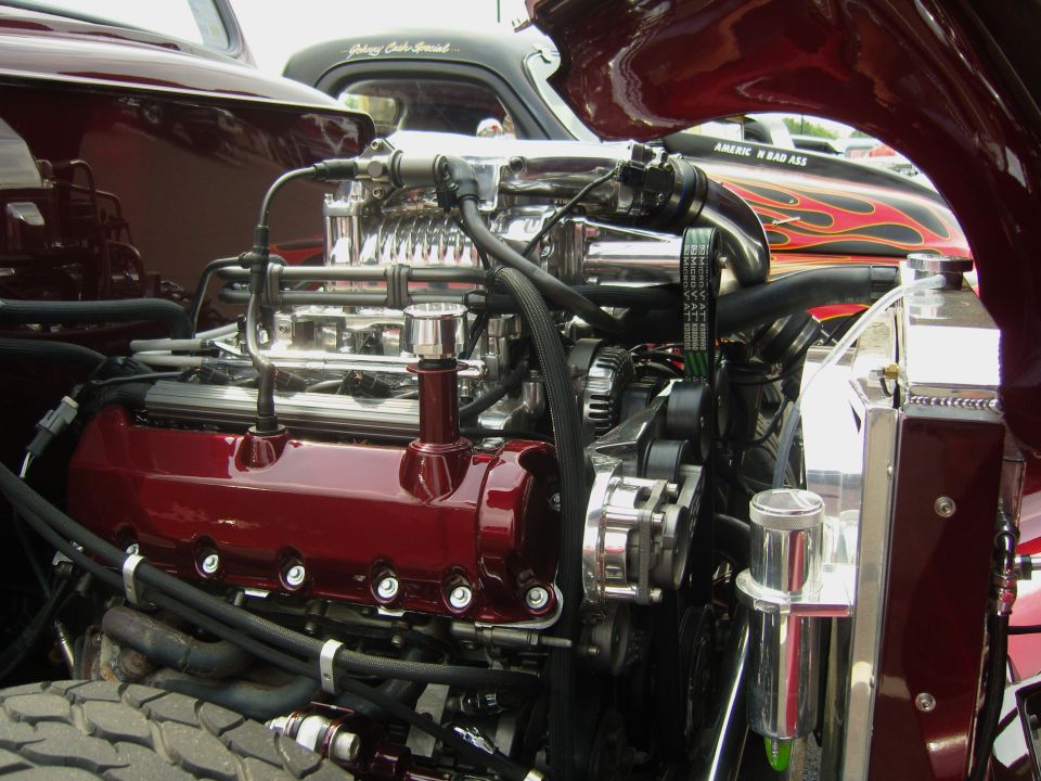 Close-up of a polished V8 engine with a supercharger, chrome components, and red valve covers. Another vehicle with flame graphics is visible behind it