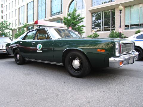 1991 Ford Crown Victoria - A 1977 Plymouth Gran Fury police car is parked on the street, showing its distinctive grille and light bar.
