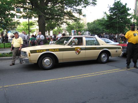 1991 Ford Crown Victoria - A tan police car with a dark green stripe and "STATE POLICE" text is parked on a street with people around.