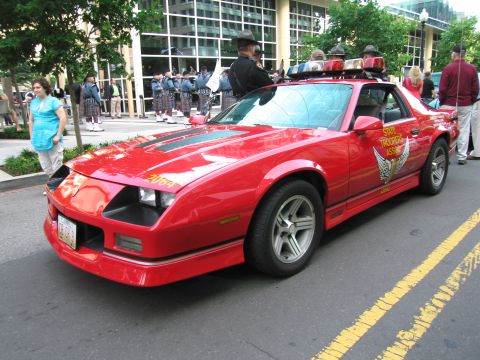 1991 Ford Crown Victoria - A red police car is parked on a street with a marching band in the background.
