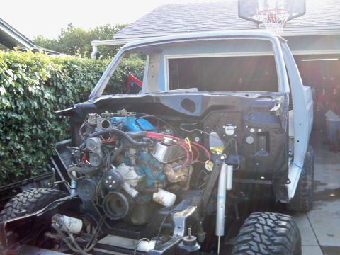 1985 Ford Bronco - A partially disassembled Ford truck sits in a driveway, with its engine and front suspension components visible.