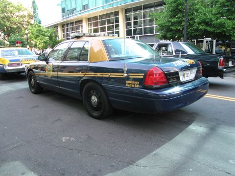 1991 Ford Crown Victoria - A dark blue police car with gold trim is parked on a street next to another vehicle.