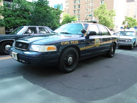 1991 Ford Crown Victoria - A dark blue police car with a yellow stripe and "STATE POLICE" text is parked on the street next to another vehicle.
