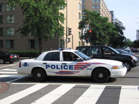 1991 Ford Crown Victoria - A white police car with red and blue stripes on its side is stopped at a crosswalk, with other vehicles visible behind it.