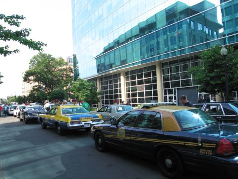 1991 Ford Crown Victoria - A line of police vehicles is parked on the street, with a large glass building reflecting the scene.