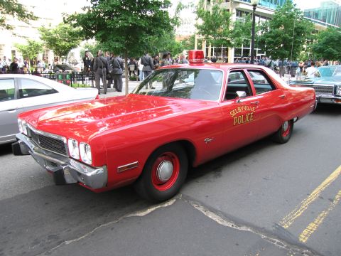 1991 Ford Crown Victoria - A vintage red police car with a light bar on top is parked on a street with other vehicles and people in the background.