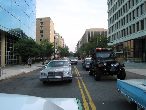 1991 Ford Crown Victoria - A convoy of vehicles, including a silver sedan and a black Jeep with emergency lights, travels down a city street.