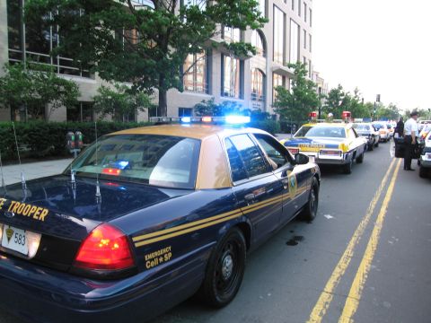1991 Ford Crown Victoria - A line of police cars, including a dark blue and tan state trooper vehicle, are parked on a city street.