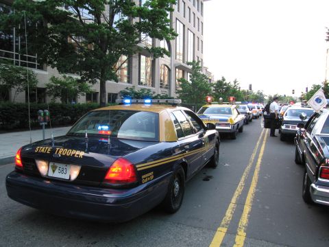 1991 Ford Crown Victoria - A line of police cars, including a dark blue and tan state trooper vehicle, are stopped on a city street.
