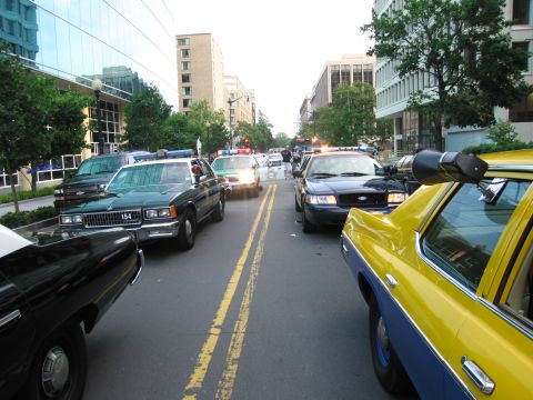 1991 Ford Crown Victoria - A line of police vehicles and a taxi are stopped in traffic on a city street with buildings on either side.