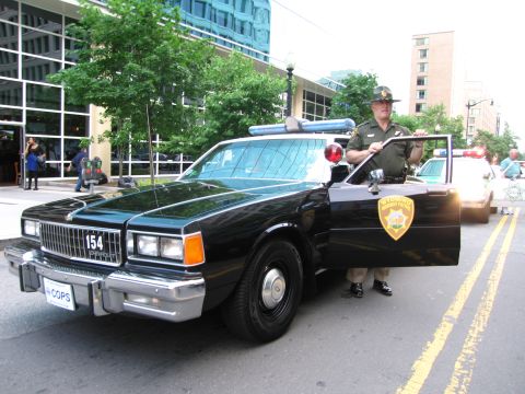 1991 Ford Crown Victoria - A police officer stands next to a black and white patrol car on a city street.