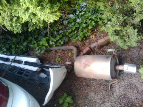 1998 Subaru Legacy - Discarded exhaust system part and a broken car bumper are visible in the yard.