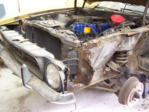 1972 Ford Mustang - The front end of a yellow car, likely from the early 1970s, shows significant rust and damage, with the hood open to reveal the engine.