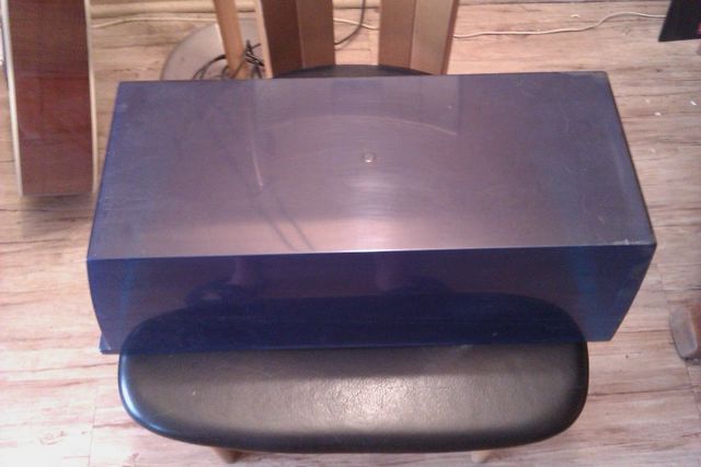 Blue electronic component sitting on a black stool, with wooden floor and speakers in background