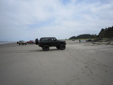 1996 Ford Bronco - A black 1996 Ford Bronco is parked on a beach, with other vehicles and people in the distance under a cloudy sky.