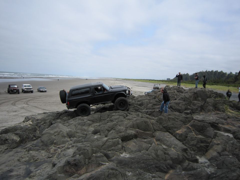 Black Ford Bronco climbing over rocks on a beach with other vehicles in the background