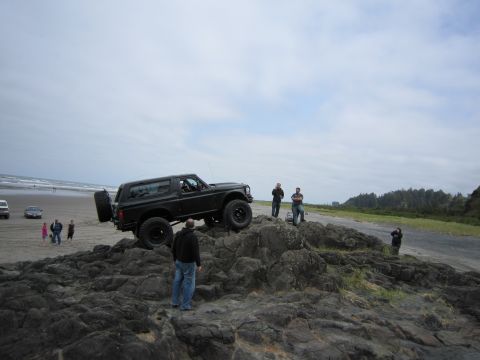 1996 Ford Bronco - A black 1996 Ford Bronco is positioned on rocks near the ocean, with several people observing.