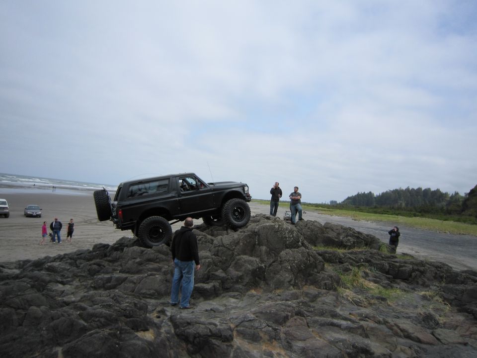 Black 1996 Ford Bronco parked on rocks near a beach with people watching